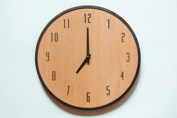 Wooden mechanical wall clock with black numerals and black hands showing seven o\'clock.