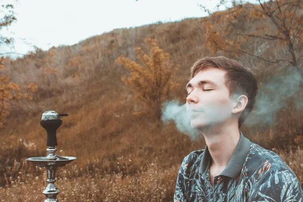 A young guy smokes a hookah in nature with his eyes closed and blows smoke from his nose.