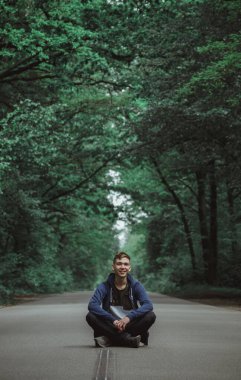 Tişörtlü genç bir adam yolun ortasında oturuyor, yoğun bir ormanla çevrili..