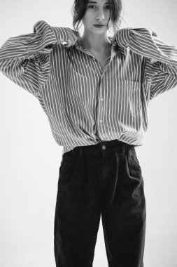 Koyu renk saçlı, kambur pantolonlu ve bol çizgili gömlekli bir kız beyaz arka planda yakasını düzeltiyor..