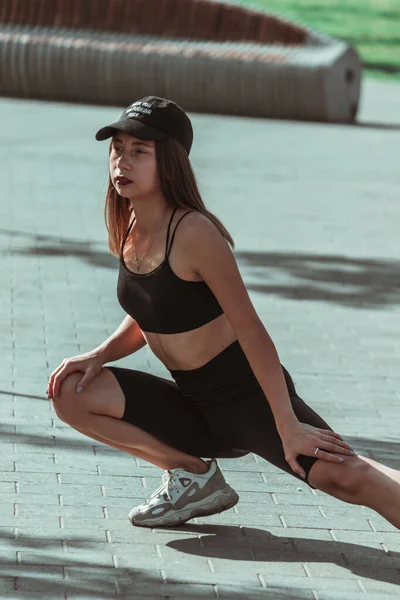 A girl in a black sports uniform and a cap is stretched out in the fresh air.