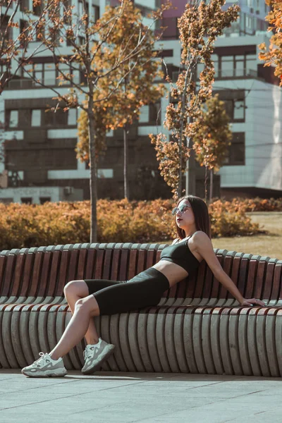 A slender girl with a toned body and abs sits on a long bench against the backdrop of a new building under the bright sun.