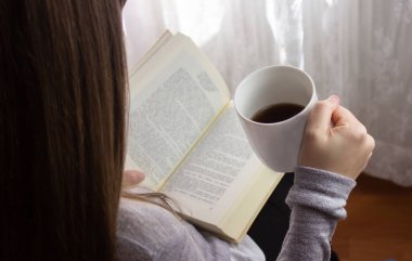 Bir fincan sıcak kahve içerken kitap okuyan bir kadın.