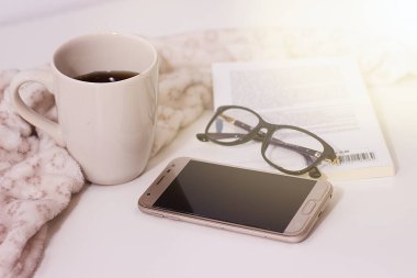 Beyaz masada bir fincan sade kahve, bir kitap, siyah bardaklar ve bir cep telefonu var..
