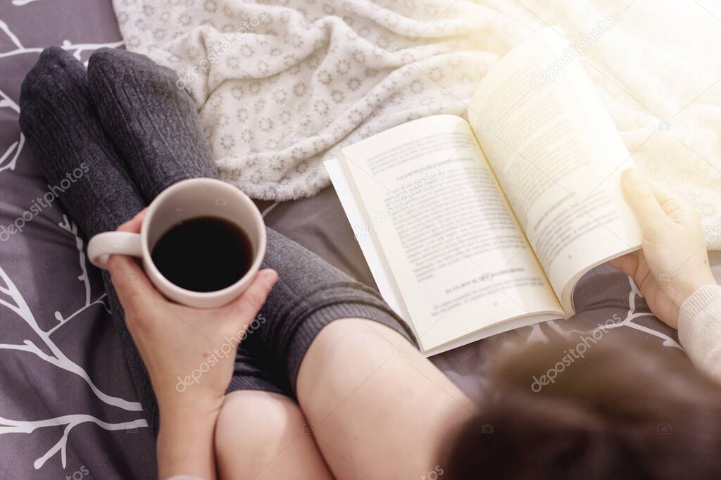 a woman drinking a black coffee and reading a book on the bed with a blanket.