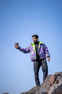 Genç Hintli yakışıklı çocuk dağlarda kar ceketi ve eşofman altı giymiş bir uçurumdan fotoğraf çekiyor..