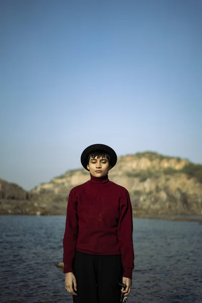 Portrait of an aesthetic looking girl wearing red sweater or sweatshirt with a black hat captured near a lake with a beautiful scenery in the background.