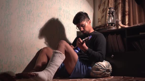 暗闇の中で骨折した足でベッドの上に座っている間 若いインド人男性はウクレレを演奏します — ストック動画
