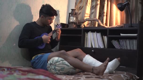 年轻的印度人坐在床上弹奏口琴 腿骨折 — 图库视频影像