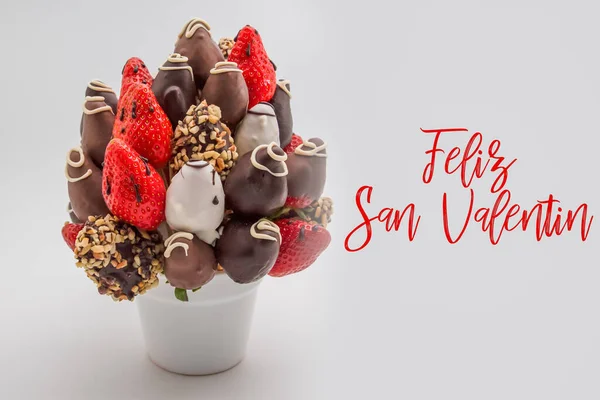 Happy valentine\'s day greeting card in Spanish with red lettering that reads Feliz San Valentin; Bunch of edible flowers, arrangement of chocolate dipped strawberries isolated on white