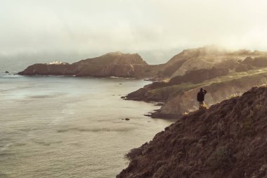 Açık havada giyinmiş bir adam okyanusun kıyısındaki bir tepede dikiliyor. Sisli bir San Francisco gününde Point Bonita Deniz Feneri 'ne bakıyor.