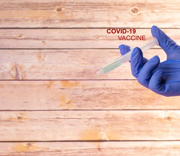 木製の背景を持つCovid 19ワクチン注射器とCovid 19ワクチンというタイトルで病院のラテックス手袋と手 — ストック写真