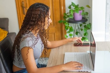 Kız, bilgisayarla uğraşırken oturma odasındaki masasında cep telefonunu kullanıyor..