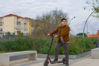 Kahverengi ceketli genç çocuk banklı bir meydanda elektrikli scooter 'a yaslanıyor.