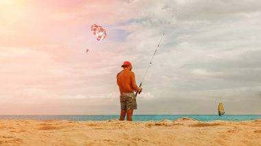 Kumsalda sarı kum ve açık mavi deniz, şortlu sıcak güneşli bir adam ve bir kep ve olta ile balıkçılık, yakınlarda bir rüzgar sörfü var, mavi gökyüzü, beyaz bulutlar turistler için bir yer.