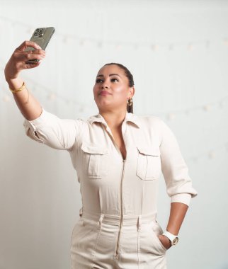 Bir kadın cep telefonuyla selfie çekiyor. O beyaz bir elbise giyiyor ve bileğinde bir saat var.