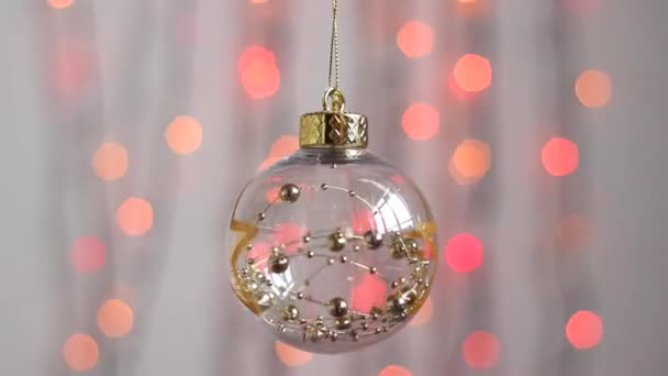 漂亮的圣诞树玩具挂在五彩缤纷的圣诞彩灯的背景上 轻轻摇曳着 透明的圣诞树玩具 形状是一个有金星和金桶的球体 — 图库视频影像