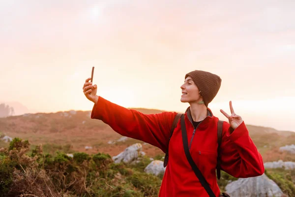 Attractive young woman in red clothes taking pictures at sunset in the field. He wears red clothes and a woolen hat. makes the symbol of victory.