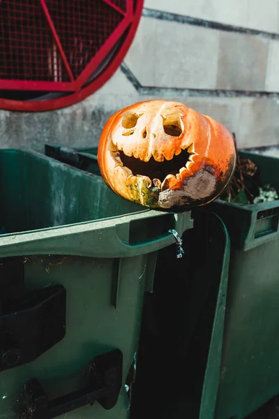 carved pumpkin with sinister smile abandoned and rotting on top of garbage container. halloween decoration. end of party.
