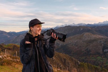 Genç fotoğrafçı gülümsüyor ve kamerayı eliyle tutarken dağlarda gün batımının güzel bir fotoğrafını çekerken bir jest yapıyor. Doğa ve boş zaman.