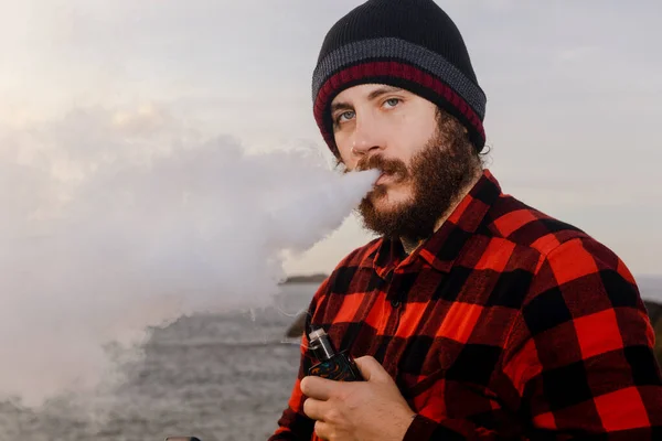 rugged looking bearded man blowing big puffs of steam while vaping. Person vaping his electronic cigarette with the sea in the background in a beautiful sunset. habits, additions and health.