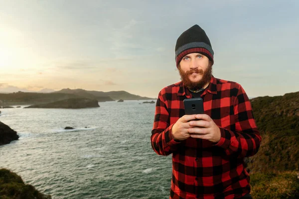 Smiling bearded man looking at camera and using his smartphone in a coastal setting.