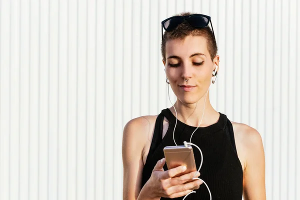 Kısa saçlı ve piercingli bir kadın sokakta akıllı telefonuyla müzik dinliyor. Sıvı ve çift cinsiyetlilik üretiyorum. Beyaz arkaplan.
