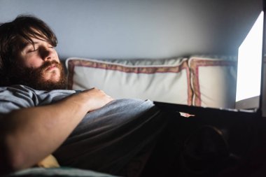 Sakallı adam kanepenin üzerinde bilgisayarla uyuyor. İnternette film izlerken uyuyakalan biri. tembellik ve yorgunluk.