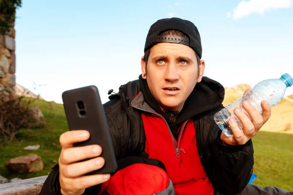 Dağlarda kaybolmuş şapkalı genç yürüyüşçü. Dağcı kendini yönlendirmek için uygulamalar kullanıyor ve elinde neredeyse boş bir su şişesi tutuyor. riskler ve dağ faaliyetleri.