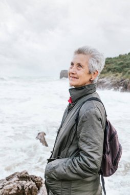 Bulutlu bir günde güçlü dalgalarla kumsalda yürürken emekliliğinin keyfini çıkaran son sınıf kadınının portresi. Emekli adam kameraya bakıyor. etkin yaşlılar.