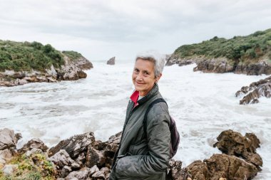 Yaşlı kadın, bulutlu bir günde güçlü dalgalarla kumsalda yürürken emekliliğinin tadını çıkarıyor. Emekli adam kameraya bakıyor. etkin yaşlılar.