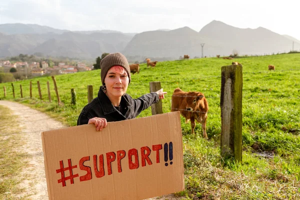 vegan animal rights activist poses with a support sign in front of livestock. young girl asking for support.