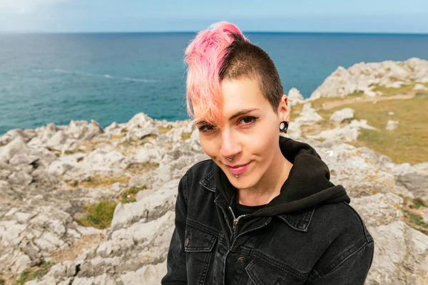 Pretty woman with punk style, pink hair and piercings looking at camera with the sea in the background. own identity. urban tribes and youth.