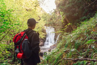 Sırt çantalı genç bir kız şelaleli bir orman manzarasını gözlemliyor. Şelalenin önündeki kadın doğanın tadını çıkarıyor. Etkin turizm.