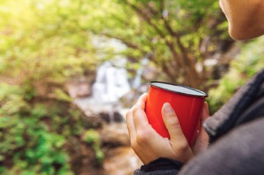 Ormandaki bir şelalenin önünde kırmızı bardağı su ile tutan tanınmayan bir yürüyüşçünün elleri. Açık hava aktiviteleri. Aktif turizm. Hafta sonu kaçamağı.
