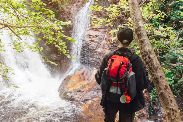 back-turned hiker with red backpack standing on a rock contemplating a waterfall in a jungle setting. Person enjoying the forest on a weekend getaway. active tourism and adventure activities.
