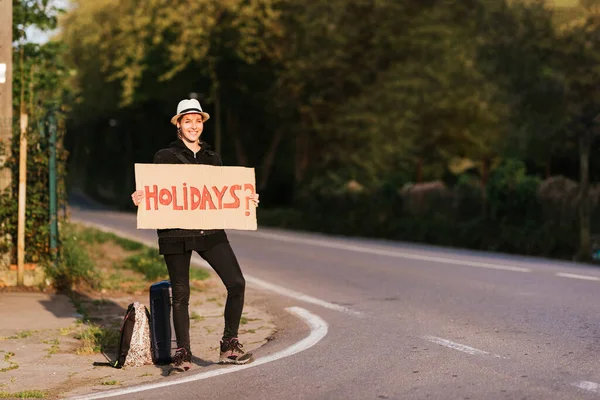 Smiling young girl doing hitchhiking on the road. standing person with hat, luggage and a sign with the word \