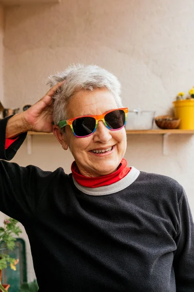 Kısa gri saçlı, gökkuşağı gururlu güneş gözlüğü takan, gülümseyen ve kafasına dokunan yaşlı lezbiyen kadının dikey portresi. İnsan hakları aktivisti. lgbt topluluğu.