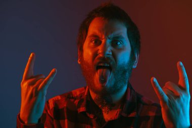 Dövmeli ve piercingli bir adam elleriyle rock ve heavy metal hareketi yapıyor. Alternatif insanlar. Boynuzları elleriyle çeken biri. Renkli ışıklı stüdyo portresi.