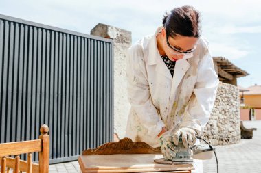 Latin kadın elektrikli zımpara makinesini ahşap mobilyaların üzerinde kullanıyor. Açık havada çalışan İspanyol biri. Küçük marangozluk ve mobilya restorasyon işi.