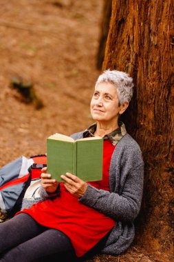 Gülümseyen ve düşünceli yaşlı bir kadın ormanda bir ağacın üzerinde kitap okuyor. Yaşlı insan doğada bir kitaptan zevk alıyor.