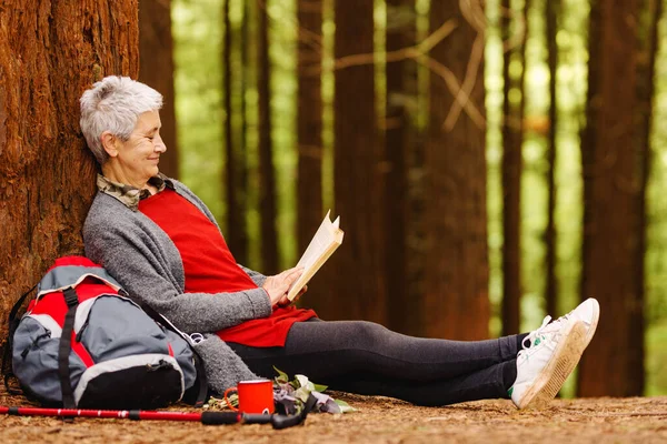 Smiling old lady with gray hair relaxes lying against a tree reading a book in the forest. senior person resting during a hike.