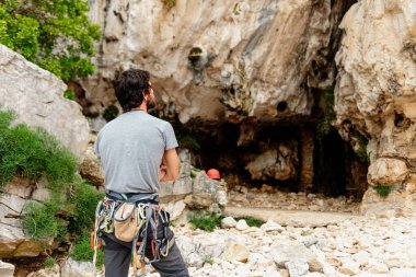Kafkasyalı genç adam kolları çapraz dönmüş ve tırmanmak için donanımlı bir şekilde kaya duvarına tırmanmış..