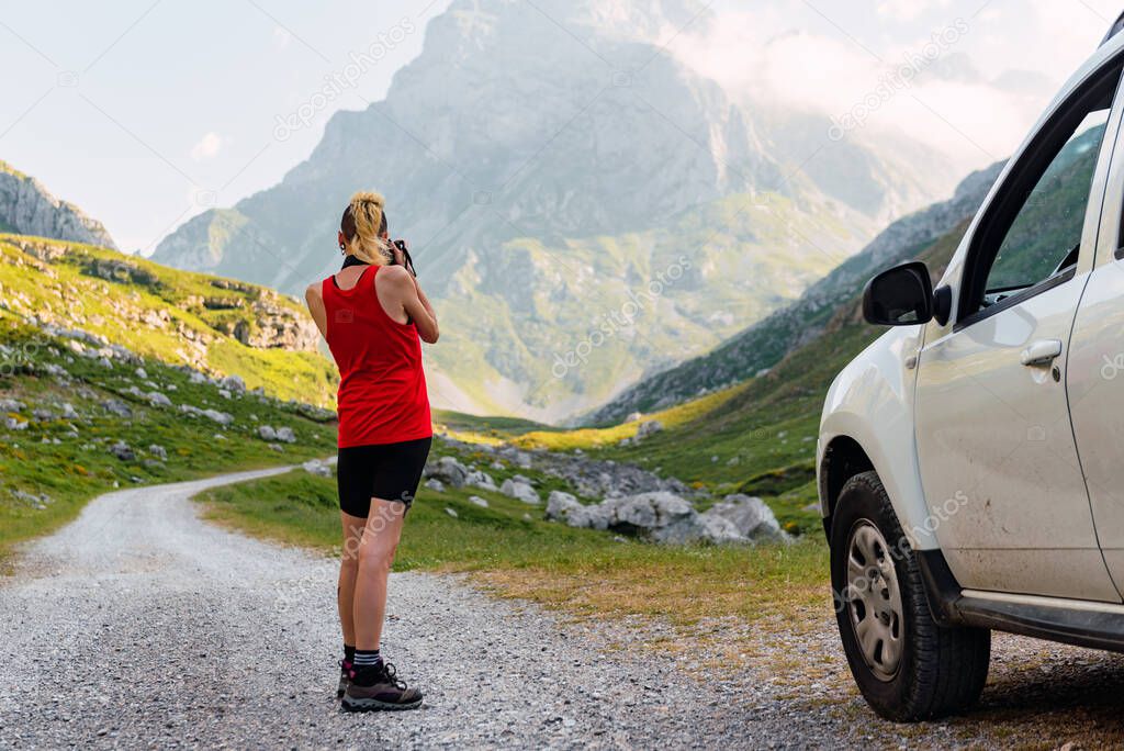 young caucasian woman in shorts taking pictures on a mountain near her white off-road car during an adventure trip.