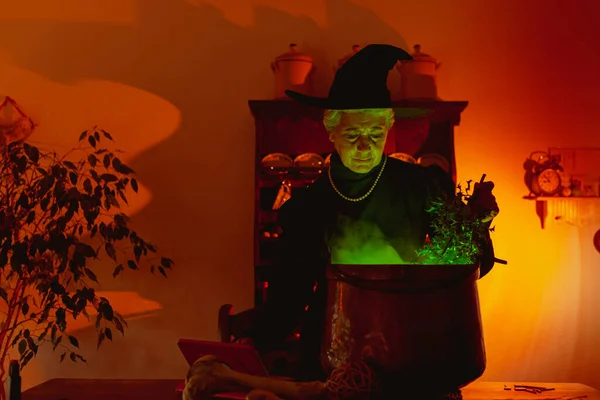 古典的な魔女として変装した年上の女性は 光と緑の煙を発する銅鍋で魔法のレシピを準備します — ストック写真
