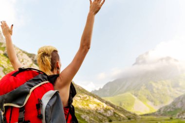 Kırmızı sırt çantalı beyaz bir kadın gün batımında bir vadi ve bir dağın önünde kollarını kaldırıyor. Hafta sonu kaçamağı ve doğada tatiller. Macera seyahatleri.