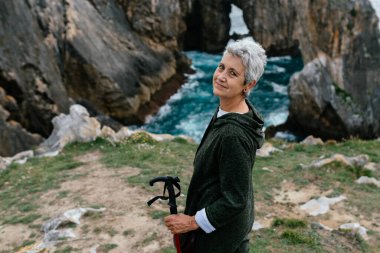 Uçurumun kenarında egzersiz yapan gri saçlı ve yürüyüş sopasıyla mutlu Kafkasyalı bir son sınıf kadını. Son sınıf öğrencisi spor yapıyor ve manzarayı inceliyor..