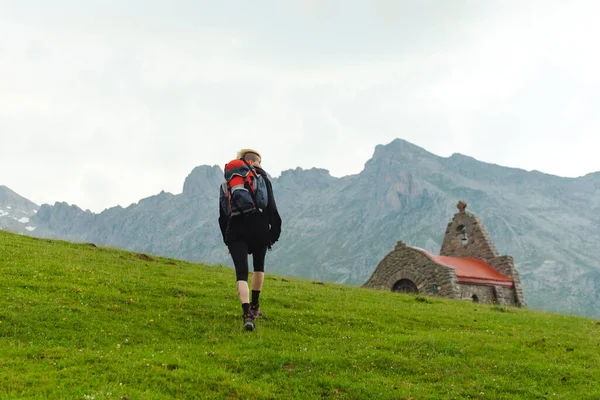 back-turned hiker with backpack hiking, ascending a green slope of the mountain valley and near a church.