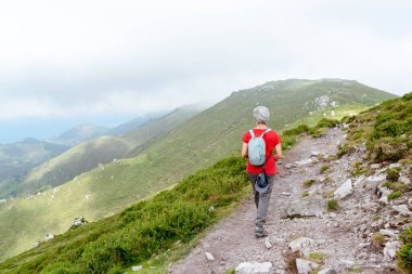 Bulutlu bir günde dağ yolunda yürüyen sırt çantalı ve gri saçlı yaşlı bir kadın yürüyüşçü.