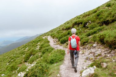 Bulutlu bir günde dağ yolunda yürüyen sırt çantalı ve gri saçlı yaşlı bir kadın yürüyüşçü.